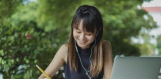 Best Essay Writer and Benefits You Can Reap Positive young Asian female student with earphones writing in copybook while doing homework at table with laptop in street cafeteria