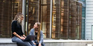 Exploring Your Options For A Home Away From Home in Australia Two women sitting on steps with a laptop, discussing ideas in urban setting.
