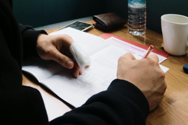 A man with a bandaged finger studies at a wooden table, accompanied by a phone and a cup.